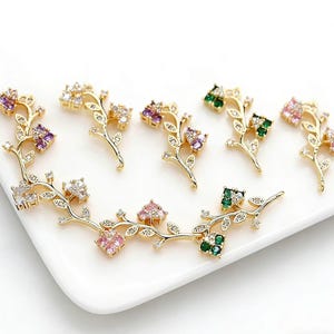 May include: Gold-toned floral jewelry components with colorful gemstone accents. The pieces feature delicate branches with leaves and clusters of purple, pink, green, and clear gemstones. The components are arranged on a white surface.