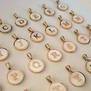 Peut inclure: Pendentifs initiaux dorés avec des centres en émail blanc. Chaque pendentif présente une lettre différente de l'alphabet, dont C, D, E, F, G, J, K, L, M, N, P, Q, R, S, T, X et Y. Les pendentifs ont une petite boucle pour une chaîne.