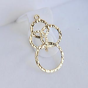 Peut inclure: Pendentif doré avec un motif de cercles entrelacés. Les cercles ont une texture martelée et sont reliés en plusieurs points. Le pendentif a une petite boucle en haut pour l'accrocher. Le fond est blanc doux.