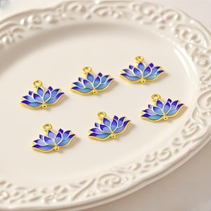 May include: Six blue and gold lotus flower charms are arranged on a white, decorative plate. The charms have a gradient of blue hues and a gold outline. Each charm has a small loop at the top for attaching to jewelry.