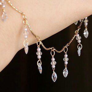 Puede incluir: Una pulsera de cadena dorada con múltiples dijes colgantes. Cada dije consta de una pequeña cuenta redonda de color lavanda pálido junto a una gota de cristal transparente facetado en forma de marquesa, suspendida por anillas doradas.