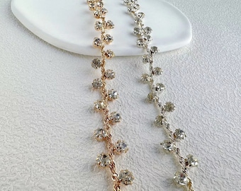Puede incluir: Dos hebras de cadena de strass, una dorada y otra plateada, con strass redondos transparentes. Las cadenas están sobre una superficie blanca.