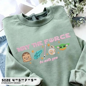 May include: A sage green crewneck sweatshirt featuring embroidered Star Wars themed graphics in pink and multi-colored thread. The design includes the text "MAY THE FORCE be with you" alongside icons of Chewbacca, a spaceship, a lightsaber, BB-8, and Grogu.