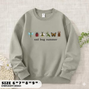 May include: A sage green crewneck sweatshirt featuring an embroidered graphic of six insects including a dragonfly, ladybug, bee, mantis, butterfly, and beetle, with the text "sad bug summer" printed in black lettering centred below the insects.