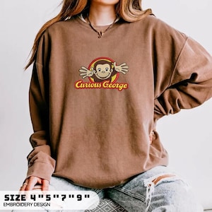 May include: A brown crewneck sweatshirt featuring an embroidered graphic of the character Curious George with arms outstretched inside a yellow and red circle. The text Curious George is embroidered in red below the character. The design is 4 by 9 inches.