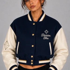 Könnte beinhalten: Eine marineblaue Collegejacke mit cremefarbenen Ärmeln, goldenen Druckknöpfen sowie geripptem Kragen, Bündchen und Saum mit weißen und goldenen Streifen. Auf der Brust befindet sich der Schriftzug Division Bleu mit einem Rauten-Symbol.