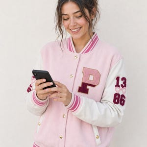 Op de afbeelding: Een lichtroze en crèmekleurig varsity-jack met een grote letter P-patch op de borst, de cijfers 13 en 86 op de mouw, en gestreepte geribbelde boorden bij de kraag, mouwen en zoom met goudkleurige drukknoopsluitingen.