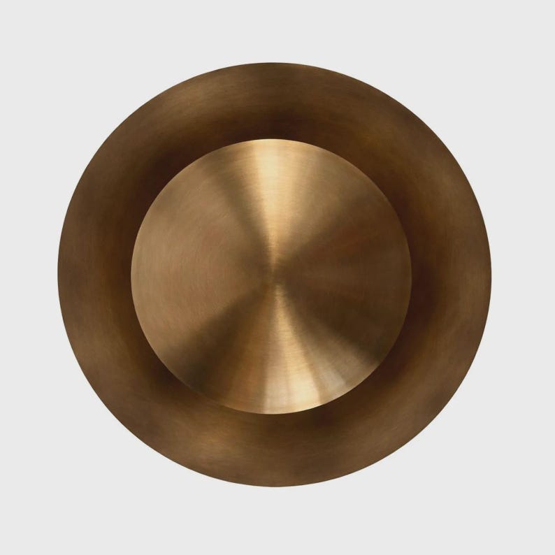 Puede incluir: Un objeto circular con un acabado en bronce met&aacute;lico, que presenta una secci&oacute;n central elevada, lisa y ligeramente abombada, rodeada por un borde exterior circular m&aacute;s ancho y plano. La superficie muestra una textura cepillada sutil.