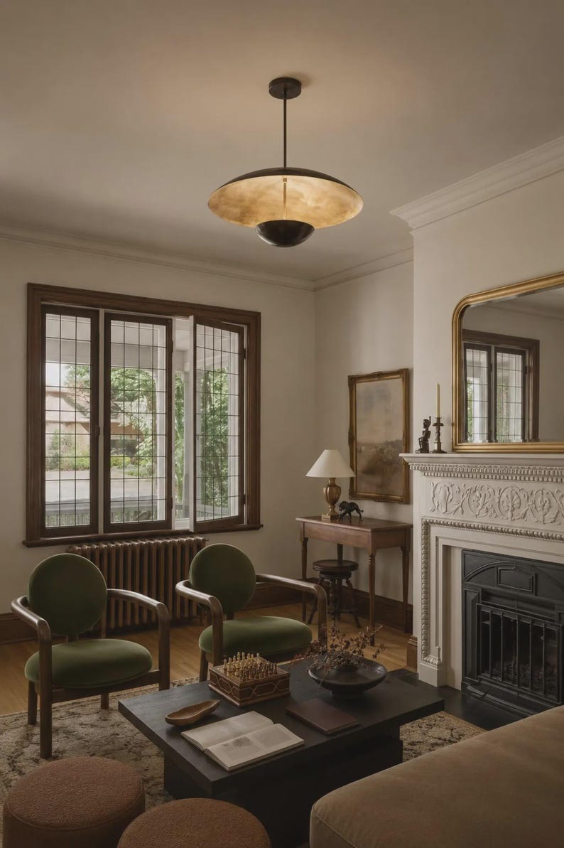 Puede incluir: Una l&aacute;mpara colgante de metal oscuro con una pantalla ancha y poco profunda en forma de platillo con un acabado interior dorado met&aacute;lico, suspendida de una varilla delgada, que ilumina hacia abajo una habitaci&oacute;n con dos sillones de terciopelo verde.