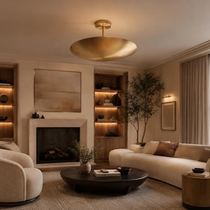 May include: A large, circular, brushed gold flush mount ceiling light fixture with a shallow, bowl-shaped shade suspended from a central stem, emitting a warm, soft glow that illuminates the surrounding living room space.