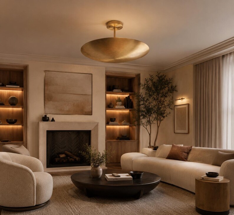 May include: A large, circular, brushed gold flush mount ceiling light fixture with a shallow, bowl-shaped shade suspended from a central stem, emitting a warm, soft glow that illuminates the surrounding living room space.