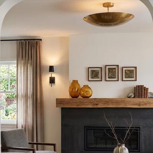 May include: A living room interior featuring a black fireplace with a thick wooden mantel, two amber glass vases, a row of books, and three framed artworks. A brass bowl ceiling light, a black wall sconce, and beige curtains are visible.