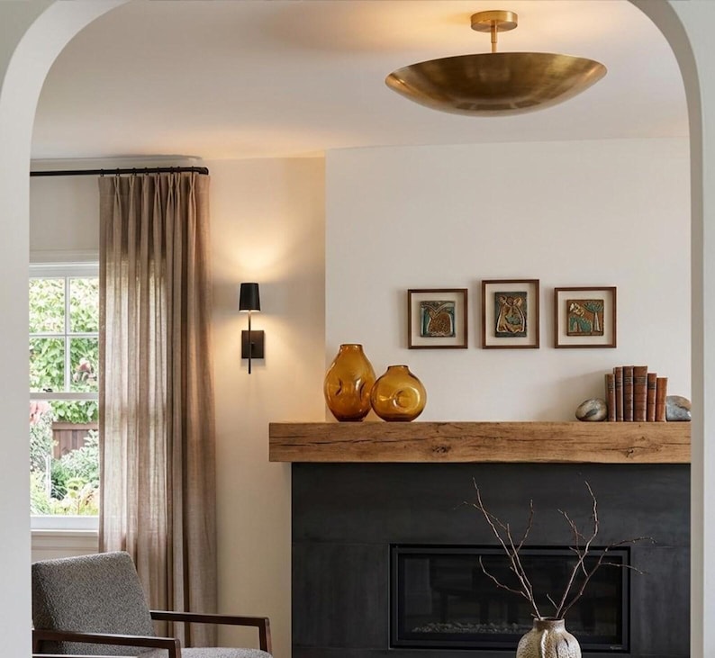 May include: A living room interior featuring a black fireplace with a thick wooden mantel, two amber glass vases, a row of books, and three framed artworks. A brass bowl ceiling light, a black wall sconce, and beige curtains are visible.