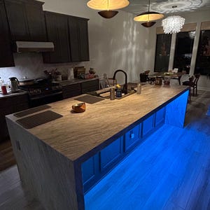 Puede incluir: Una isla de cocina con encimera de madera clara y armarios oscuros. La base de la isla cuenta con una vibrante iluminaci&oacute;n LED azul. Sobre la isla cuelgan dos l&aacute;mparas colgantes modernas con pantallas anchas y poco profundas.