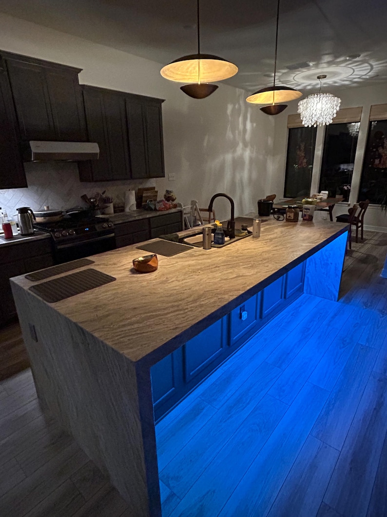 Puede incluir: Una isla de cocina con encimera de madera clara y armarios oscuros. La base de la isla cuenta con una vibrante iluminaci&oacute;n LED azul. Sobre la isla cuelgan dos l&aacute;mparas colgantes modernas con pantallas anchas y poco profundas.