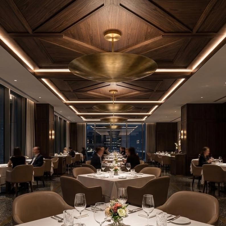 May include: A restaurant interior featuring a series of large, circular, brushed brass pendant lights suspended from a dark wood coffered ceiling with integrated linear lighting. The dining room is furnished with white-clothed tables and chairs.