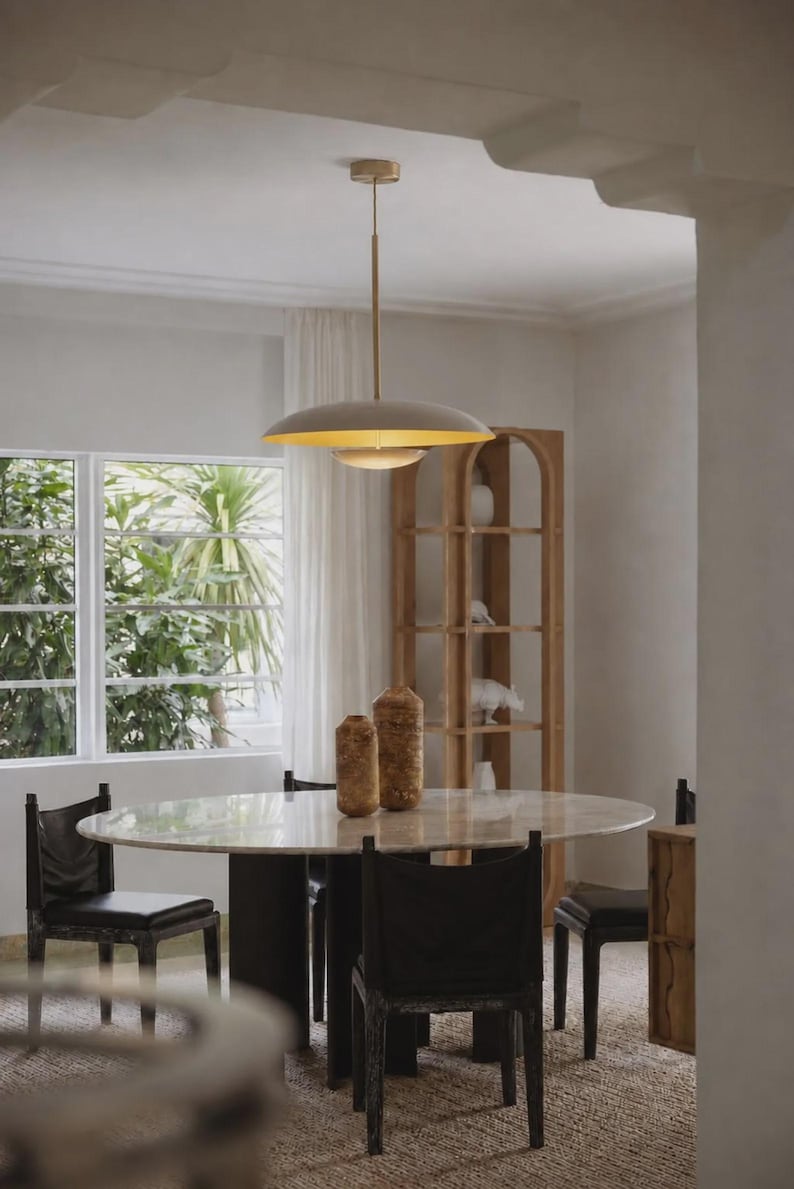 Puede incluir: Una l&aacute;mpara colgante con una pantalla de c&uacute;pula ancha y poco profunda en color beige mate y un interior dorado, suspendida por una varilla delgada desde un dosel de techo a juego. La luz emite un brillo c&aacute;lido hacia abajo.