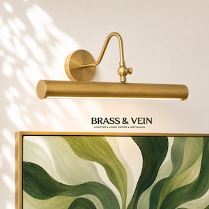 May include: A brass wall-mounted picture light with a cylindrical shade and an adjustable arm. The fixture is positioned above a painting with green abstract patterns. The text BRASS & VEIN LIGHTING HOME DECOR ARTISANAL is visible below.