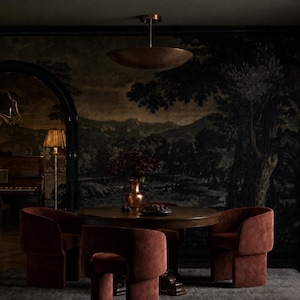 May include: A round wooden dining table surrounded by four rust-colored upholstered chairs with curved backrests. A copper-toned bowl sits on the table next to a vase. A large copper pendant light hangs above, and a floor lamp stands nearby.