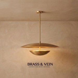 May include: A brass pendant light featuring a wide, flat circular shade positioned above a smaller, bowl-shaped shade, suspended by a thin vertical rod. The text BRASS & VEIN LIGHTING • HOME DECOR • ARTISANAL is printed in black below the fixture.