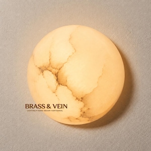 Op de afbeelding: Een ronde wandlamp met een doorschijnend, warm gekleurd stenen oppervlak met natuurlijke aders die oplichten bij verlichting. De tekst BRASS & VEIN LIGHTING HOME DECOR ARTISANAL staat in donkerbruine letters aan de linkeronderzijde.