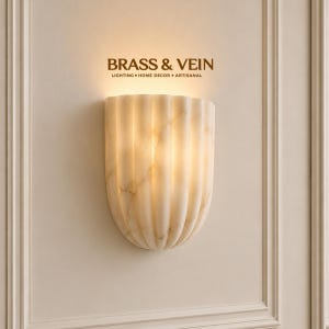 Könnte beinhalten: Eine halbkreisförmige Wandleuchte aus cremeweißem Marmor mit vertikalen Rillen. Die Leuchte strahlt nach oben ein warmes, sanftes Licht aus. Über der Leuchte ist der Text BRASS & VEIN LIGHTING • HOME DECOR • ARTISANAL in Braun aufgedruckt.