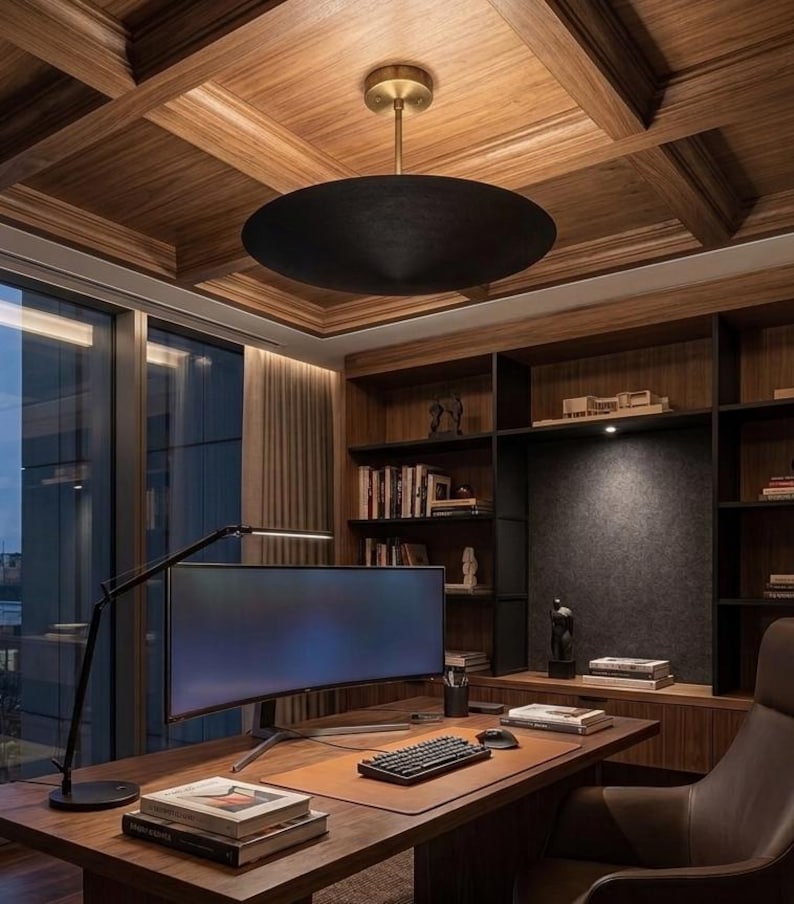 May include: A large, matte black, disc-shaped pendant light fixture with a brass-toned ceiling mount hangs above a wooden desk. The desk features a curved computer monitor, a black keyboard, a computer mouse, and stacks of books.