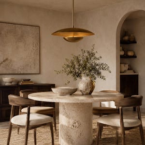 Puede incluir: Una mesa de comedor redonda con una base de pedestal de piedra texturizada y una superficie lisa, rodeada por cuatro sillas de madera con asientos tapizados en tonos claros. Una gran l&aacute;mpara colgante de metal dorado cuelga arriba.