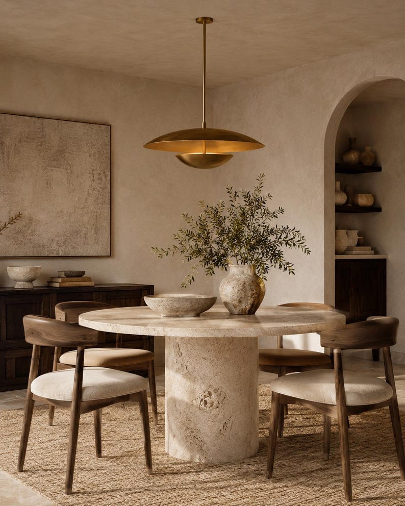 Puede incluir: Una mesa de comedor redonda con una base de pedestal de piedra texturizada y una superficie lisa, rodeada por cuatro sillas de madera con asientos tapizados en tonos claros. Una gran l&aacute;mpara colgante de metal dorado cuelga arriba.