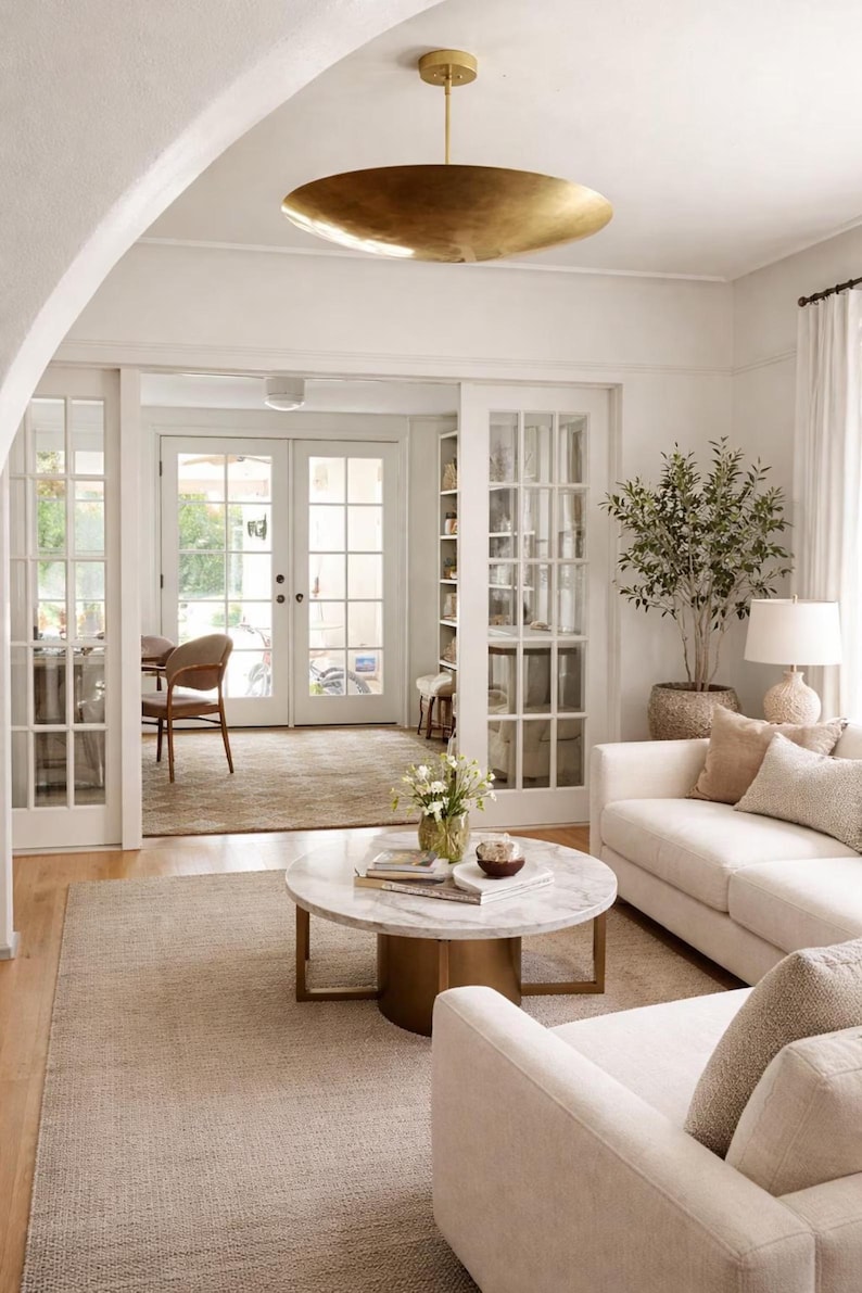 May include: A living room featuring a round white marble coffee table with a gold base, a cream-colored sofa with textured throw pillows, a large brass pendant light, a potted olive tree, and white French doors leading to an adjacent room.