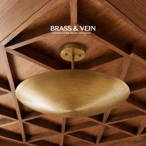 May include: A brushed brass flush mount ceiling light fixture with a wide, shallow, disc-shaped shade. The fixture is mounted to a wooden ceiling. The text BRASS & VEIN LIGHTING HOME DECOR ARTISANAL is visible above the light fixture.