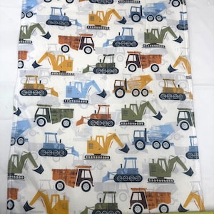 May include: A white fabric featuring a repeating pattern of construction vehicles including yellow, blue, green, and orange dump trucks and excavators. A yellow measuring tape is positioned at the bottom, showing a width of approximately 15 inches.