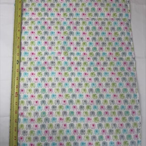 May include: A rectangular fabric piece featuring a repeating pattern of small elephants in pink, blue, green, and gray on a white background. A yellow measuring tape is placed along the left edge, showing a length of approximately 24 inches.