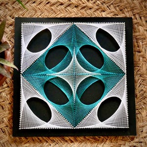 Op de afbeelding: Een vierkant draadkunstwerk op een zwart bord met een geometrisch patroon gemaakt van witte en blauwgroene draden. Het ontwerp bestaat uit elkaar kruisende curven en lijnen die een centrale ruitvorm vormen met vier omliggende ovale openingen.