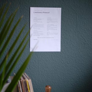 Könnte beinhalten: Ein weißer, rechteckiger Papierausdruck mit dem Titel Consistency Protocol ist an einer dunkel-petrolfarbenen, strukturierten Wand befestigt. Das Dokument enthält schwarzen Text, der in Abschnitte wie Purpose, Process, Discipline, Execution, Mental Framework und Review unterteilt ist.