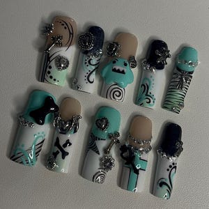 Könnte beinhalten: Ein Set aus zehn künstlichen Fingernägeln in den Farben Minzgrün, Schwarz und Nude. Die Designs umfassen handgemalte Wirbel, Streifen und Kreuzmotive, akzentuiert durch 3D-Anhänger wie Metallherzen, Sterne, Blumen und ein kleines blaues Monster.