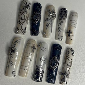 Könnte beinhalten: Ein Set aus zehn künstlichen Fingernägeln in Schwarz und Weiß. Die Designs umfassen komplexe Schnörkel, handschriftlichen Text und strukturierte schwarze Muster. Mehrere Nägel sind mit silberfarbenen Kreuz-Anhängern und kleinen Strasssteinen verziert.