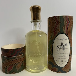 May include: A clear glass bottle of Ralph Lauren Thoroughbred home fragrance spray with a gold cap, accompanied by a cylindrical cardboard container featuring a paisley pattern and a label depicting a person on a horse, marked 5.7 fl. oz.