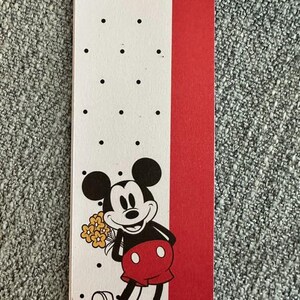 May include: A rectangular paper bookmark featuring a white section with black polka dots and a solid red vertical stripe. The design includes a graphic of Mickey Mouse holding a small bouquet of yellow flowers, wearing red shorts and white shoes.