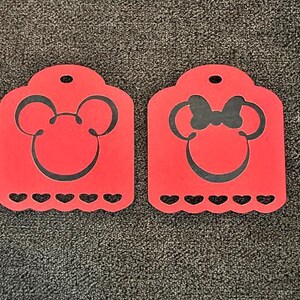 May include: Two red rectangular tags with rounded tops and scalloped bottoms featuring heart-shaped cutouts. The left tag displays a black silhouette of a mouse head, while the right tag features a black silhouette of a mouse head with a bow.