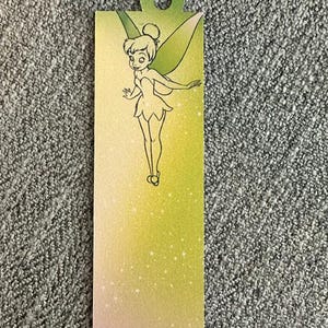 May include: A rectangular bookmark featuring a gradient background that transitions from a soft pink at the bottom to a vibrant lime green at the top. A black line art illustration of a fairy with wings is centered, and small white stars are scattered.
