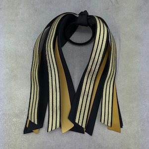 May include: Black and gold striped ribbon hair tie. The ribbon is tied in a bow with the ends of the ribbon hanging down.