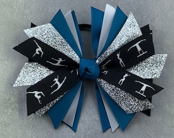 May include: A teal, silver and black hair bow with a white gymnast silhouette pattern. The bow is tied with a teal elastic band.