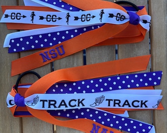 May include: Two hair ties with layered ribbons in orange, white, and purple. One ribbon features the text NSU, while the other displays NALA. Additional ribbons show white polka dots on purple, and white ribbons with black graphics of runners, arrows, and the word TRACK.