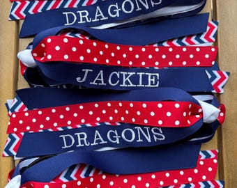 May include: A collection of layered hair bows made from grosgrain ribbon in navy blue, white, and red. The ribbons feature polka dot and chevron patterns. Each bow is embroidered with white text reading DRAGONS, JACKIE, or ELIZA.
