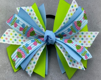 May include: A blue and green hair bow with a fish print. The bow is made of multiple layers of ribbon and is tied with a green elastic band.