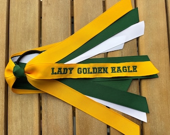 May include: A hair accessory featuring multiple long ribbons in yellow, dark green, and white attached to a black elastic hair tie. The top yellow ribbon is embroidered with the dark green text LADY GOLDEN EAGLE in a bold, capitalised font.