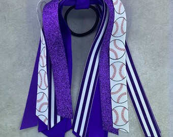 May include: Purple and white hair tie with baseball print ribbon. The hair tie has a purple glitter ribbon, a white ribbon with purple stripes, and a white ribbon with baseball print.