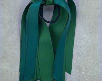 May include: Green and teal ribbon hair tie with a bow. The ribbon is tied in a knot and the ends are left long.