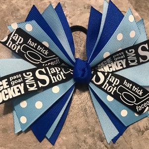 May include: A blue and white polka dot hair bow with black ribbon featuring hockey-themed text, including "Slap Shot", "Hat Trick", "Faceoff", "Pass Goal", and "Ice Hockey".